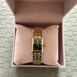 Guldfärgad HIMO klocka med svart urtavla - Stilren klocka från HIMO med rektangulär svart urtavla och romerska siffror. Armbandet är guldfärgat och har en klassisk länkdesign. Perfekt accessoar för dig som gillar en elegant och tidlös look. Kostade 1399 kr är oanvänd .,  