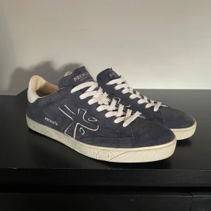 Premiata Skor - Snygga sneakers från Premiata i mörkblå mocka med vita detaljer och snörning. Skorna har en vit sula med tryck och en kontrasterande vit hälkappa med logga. Perfekta för dig som gillar stilrena och trendiga sneakers med premiumkänsla. Storlek 43, köpta på farfetch, box finns aldrig använda endast testade såklart. Öppen för prisförslag.