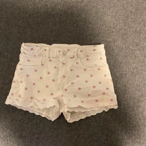 Vita blommiga shorts med spetskant - Söta vita shorts med små rosa blommor och spetskant nertill. Högmidjade modell med klassiska fickor fram och bak, samt dragkedja och knapp. Perfekta för sommarens varma dagar och ger en gullig vibe till din outfit. Storlek 8-9. Använda. 