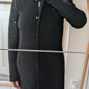 Svart kappa från ONLY med huva XS - Säljer en svart, lång och stilren kappa från ONLY i storlek XS. Jackan har huva, dragkedja och knappar framtill samt slät insida och med öppna fickor på sidorna. Perfekt för dig som gillar en klassisk look med enkel design och snygg passform. Kan vara hårig/katter förekommer i hemmet och därav säljer jag den. Håret går att tvätta bort eller använda en roller.