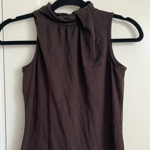 Brun ärmlös topp med knyt från Esprit - Snygg brun ärmlös topp från Esprit med hög hals och knyt-detalj vid kragen. Tillverkad i mjuk bomull, perfekt för en stilren och trendig look. Passar dig som gillar minimalistiska plagg med en twist.