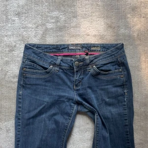 Blå bootcut jeans från ONLY W29 L32 - Snygga blå jeans från ONLY med klassisk femficksdesign och bootcut-ben. Jeansen har normal passform, låg midja och är tillverkade i mjukt bomullsmaterial med lite stretch. Fina detaljer med nitar på bakfickorna och rosa insida i linningen.