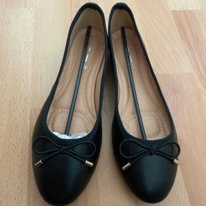 Helt nya ballerinaskor med rosett i storlek 40. - Stilrena svarta ballerinaskor med dekorativ rosett framtill med guldfärgade detaljer. Skorna har en platt sula och är gjorda i syntetmaterial med mjuk innersula för extra komfort. Perfekta för en enkel och klassisk look. Jag står inte för retur eller för postens slarv. 