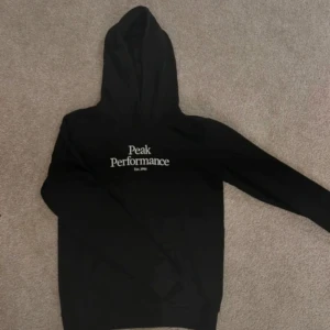 Svart hoodie från Peak Performance - Svart hoodie från Peak Performance med vit logga på bröstet. Klassisk modell med luva och långa ärmar. Oanvänd. 