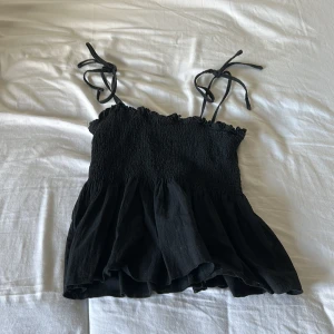 Svart smockad linnetopp från SHEIN - Supersöt svart linnetopp från SHEIN med smockad överdel och volangig nederdel. Axelbanden är tunna och knyts på axlarna för en extra gullig touch. Perfekt till jeans eller kjol när du vill ha en chill men ändå snygg look.