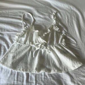 Vit volangig linnetopp med knyt - Supersöt vit linnetopp med smala axelband, volangdetaljer längs urringningen och en liten knytning framtill. Toppen har en lätt skrynklig struktur och är luftig med en flowy passform, perfekt för varma dagar.