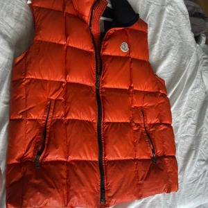 Orange dunväst från Moncler - Säljer en riktigt snygg orange dunväst från Moncler med hög krage och två dragkedjefickor. Västen har quiltad design och klassisk Moncler-logga på bröstet. Perfekt att slänga över en hoodie för en cool look.