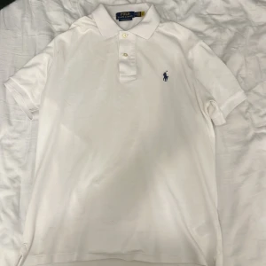 Vit pikétröja Polo Ralph Lauren  - Klassisk vit pikétröja från Polo Ralph Lauren i bomull. Tröjan har korta ärmar, krage och knäppning framtill samt den ikoniska broderade loggan i mörkblått på bröstet. Modellen är custom slim fit för en snyggare passform.