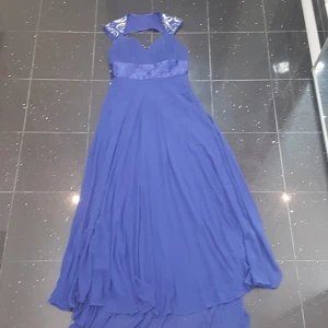 Blå långklänning med paljett-detaljer - Elegant blå långklänning med v-ringad överdel, plisserad midja i satin och axelparti dekorerat med silvriga paljetter i swirl-mönster. Klänningen har dragkedja i sidan och ett flytande, svepande tyg som ger en lyxig känsla.