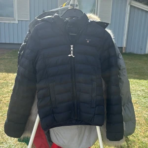 Svart dunjacka från GANT - Snygg svart dunjacka från GANT med broderad logga på bröstet. Jackan har quiltad design, dragkedja framtill och huva. Perfekt för kyliga dagar och enkel att matcha med olika outfits. Nypris 1500kr.