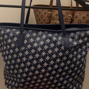 Blå DON DONNA väska - Snygg blå shoppingväska från Don donna med vitt monogrammönster över hela väskan. Rymlig modell med långa handtag, perfekt för att bära allt du behöver. Klassisk och stilren design som lyfter vilken outfit som helst.