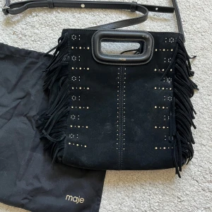 Maje väska - Snygg svart axelväska i mocka från Maje med coola fransar på sidorna och små nitar i mönster framtill. Väskan har både handtag och justerbar axelrem, perfekt för att lyfta din outfit. Kommer med original dustbag.