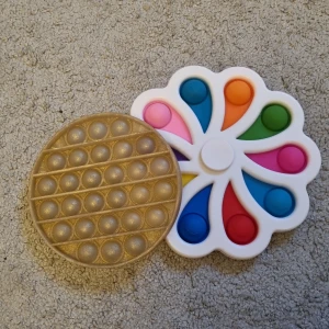 En Stor Fidget Spinner med Bubblor +En Guldig Pop It Fidget Toy  - Två olika pop it-fidgetleksaker, en rund i guld och en blomformad med färgglada bubblor. Perfekt för stresshantering, avslappning eller bara för att ha kul! Passar både ungdomar och unga vuxna som gillar taktila prylar och vill ha något att pilla med.