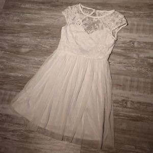 Vit spetsklänning från NLY One - Superfin vit kortklänning från NLY One med spets upptill och blommiga broderier. Kjolen är i flera lager tunn tyll som ger en luftig känsla. Korta ärmar, knappar i nacken och hjärtformad urringning. Perfekt för student eller sommarens alla tillfällen! Resårfästet på en knapp i nacken har lossnat men lätt att fixa eller bara ta bort.