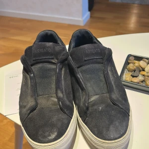Svarta sneakers från Axel Arigato - Stilrena navy blue från Axel Arigato i mocka med diskret logga i guld på sidan. Skorna har en vit platt sula och minimalistisk design utan synlig snörning, vilket ger en clean look. Perfekta för dig som gillar enkel och modern stil. St 42 men funkar även bra för 43. PRIS kan självklart diskuteras. Smutset går bort väldigt enkelt bara att borsta bort det 