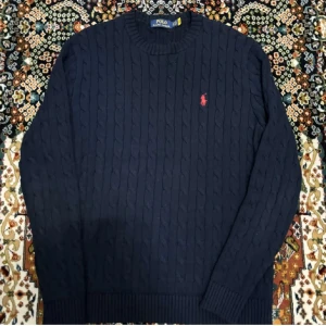Mörkblå stickad tröja från Polo Ralph Lauren - Kabelstickad mörkblå tröja från Polo Ralph Lauren med rund halsringning och klassisk röd broderad logga på bröstet. Tröjan har långa ärmar och ribbade muddar för en snygg passform. Perfekt för dig som gillar stilren och tidlös design.