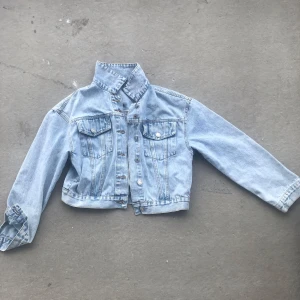 Ljusblå croppad jeansjacka - Snygg ljusblå jeansjacka i croppad modell med klassiska bröstfickor och silvriga knappar. Jackan har lång ärm och markerad krage, perfekt för en chill och trendig look. Passar till allt och ger en cool vibe till outfiten.