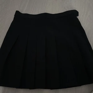 Svart plisserad kortkjol - Snygg svart plisserad kortkjol med två knappar i midjan som väljs baserat på storleksbehov därmed passar den  XXS, XS och S.  Som ny