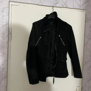 Svart jacka med dragkedjor från H&M - Svart jacka från H&M med coola silverfärgade dragkedjor på bröstet och knappar framtill. Jackan har hög krage, lång ärm och en figurnära passform. Materialet är mjukt och lätt, perfekt för dig som gillar en stilren look med edge.