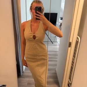 Beige långklänning med halterneck - Snygg beige långklänning med halterneck och öppen rygg. Klänningen har en liten cut-out framtill med metallspänne och figurnära passform. Perfekt för sommarens alla tillfällen när du vill känna dig extra chic.