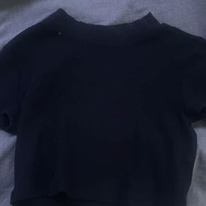 Svart croppad ribbad topp - En svart croppad topp med korta ärmar och ribbad struktur. Toppens material är stretchigt och mjukt, perfekt för en chill och stilren look. Enkel design utan synliga detaljer eller mönster, passar till allt.