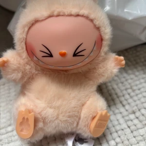 Pop Mart beige fluffig labubu  - Supersöt beige labubu från Pop Mart i fluffigt syntetmaterial. Labubun har ett gulligt ansikte med stort leende och öron, samt en praktisk rem med Pop Mart-logga. Perfekt för dig som vill ha något unikt och lekfullt till din outfit.