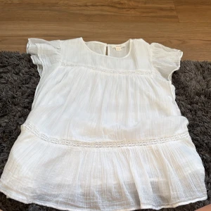 Vit blus med volangärmar - Luftig vit blus i tunn bomull med volangärmar och fina spetsdetaljer. Blusen har rund halsringning och ett lätt rynkat fall som ger en romantisk vibe. Perfekt för varma dagar när du vill ha något svalt och stilrent.