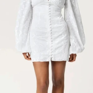 Adoreklänning  - Superfin vit kort klänning med broderade blommor, puffiga långärmar och knappar framtill. Klänningen har v-ringning och markerad midja som ger en snygg siluett. Perfekt för sommarens alla tillfällen! Klänningen är använd vid ett fåtal tillfällen och är köpt från bubbelroom för 600kr men är nu helt slutsåld. ❤️