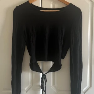 Svart croppad långärmad topp med knyt - Säljer en svart långärmad croppad topp med snygg knytning baktill. Toppen har rund halsringning och är tillverkad i mjukt bomullstyg. Perfekt för dig som gillar trendiga och stilrena plagg med en twist.