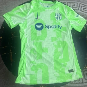 Barcelona Nike fotbollströja neon - Säljer en FC Barcelona fotbollströja från Nike i neonljusgrön färg med blå detaljer. Tröjan har klubbmärke, Nike-logga och Spotify-tryck på bröstet samt UNHCR-logga på ryggen. Tillverkad i lätt och ventilerande funktionsmaterial, perfekt för match eller träning.