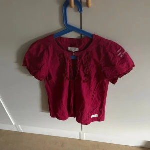 Rosa blus från Odd Molly med volang - Supersöt rosa blus från Odd Molly med korta puffärmar och volangdetaljer längs knäppningen framtill. Blusen har rund halsringning, liten knapp och broderad text på ena ärmen. Perfekt för dig som gillar färg och detaljer.