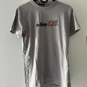 Grå Nike Air t-shirt - Snygg grå t-shirt från Nike med klassiskt Air-tryck på bröstet och orange swoosh-detalj. T-shirten har rund hals och korta ärmar, perfekt för en sportig och avslappnad stil. Tillverkad i mjuk bomull som känns skön mot huden.