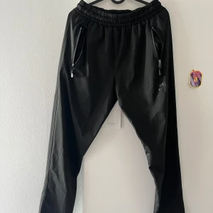 Svarta Nike Air byxor med dragkedjor - Svarta byxor från Nike Air med elastisk midja och dragkedjor på fickorna fram och bak. Snygg sportig look med diskret logga på benet. Perfekta för chill eller träning, tillverkade i ett lätt och smidigt material.