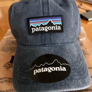 Blå keps från Patagonia - Säljer en snygg blå keps från Patagonia med klassisk logga framtill i brodyr. Kepsen har böjd skärm och justerbar rem baktill. Tillverkad av återvunnen ull och nylon, perfekt för dig som gillar stilren och hållbar streetwear. Kepsen är helt ny MED PRISLAPP.