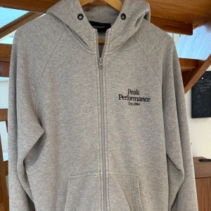 Grå hoodie från Peak Performance - Snygg ljusgrå hoodie från Peak Performance med dragkedja och logga på bröstet, sparsamt andvänd och i fint skick, nypris ligger på 1399kr. Storlek L
