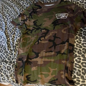 Camo t-shirt från Vans - Säljer en kortärmad t-shirt från Vans med klassiskt camouflagemönster i grönt, brunt och svart. Stor Vans-logga med skateboard på bröstet och ryggen. Skön passform och perfekt för dig som gillar streetwear.