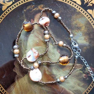 Brunt pärlhalsband med snäckdetaljer - Unikt halsband med små bruna glaspärlor, skimrande guld- och silverfärgade pärlor samt oregelbundna snäckbitar i brunt. Några större bärnstensfärgade pärlor ger extra wow-effekt. Justerbart med silverfärgad kedja och lås.