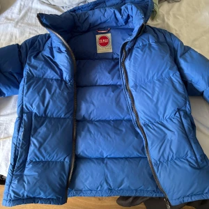 Blå Colmar pufferjacka med huva Stl M - Säljer en blå pufferjacka från Colmar med huva och dragkedja framtill eftersom jag inte använder den längre. Inga hål och är som ny. Modellen på bilden är 181 cm. Jackan har en clean look med diskret logga på ärmen och är perfekt för kalla dagar. Fylld med dun och har långa ärmar. 