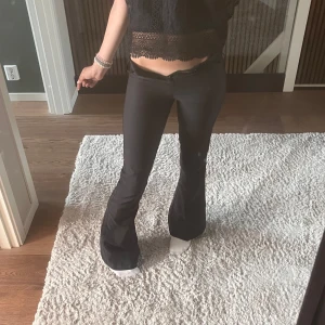 Svarta bootcut byxor med fickdetalj - Säljer ett par svarta bootcut byxor med låg midja och snygg ficka med knappdetalj bak. Byxorna har en tight passform upptill och vida ben nertill. Perfekta för dig som gillar en trendig siluett och vill ha något som sticker ut lite extra.