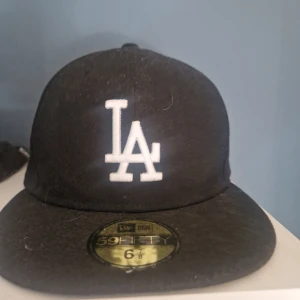 Svart LA New Era 59FIFTY keps - Klassisk svart New Era 59FIFTY keps med LA-logga broderad i vitt framtill och New Era-logga på sidan. Flat skärm och strukturerad passform. Perfekt för dig som gillar streetwear och sportig stil.