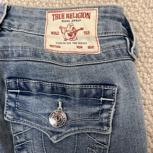 True Religion Becca Bootcut Jeans - Säljer ett par ljusblå True Religion Becca jeans med bootcut och snygga detaljerade bakfickor med knappar. Jeansen har midwaist passform och klassisk denimlook med kontrastsömmar. Perfekta för dig som gillar en tidlös och avslappnad stil.