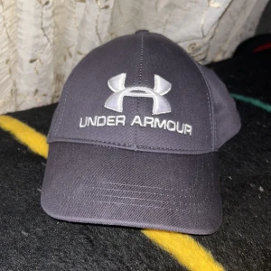 Svart keps från Under Armour - Snygg svart keps från Under Armour med broderad logga och text framtill samt baktill. Justerbar rem baktill för perfekt passform. Klassisk böjd skärm och stilren design, perfekt för en sportig look.