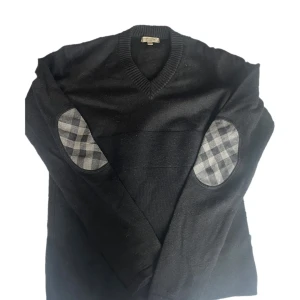 Svart Burberry stickad tröja M - Svart stickad tröja från Burberry med v-ringning och rutiga armbågslappar i grå toner. Tröjan är i ull och har långa ärmar. Perfekt för dig som gillar klassisk stil med en twist. Tillverkad i Italien. Riktigt snygg och lyxig tröja! 