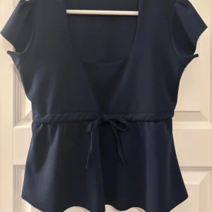 Marinblå topp med knytning från Missay - Supersnygg marinblå topp från Missay med korta ärmar och rundad urringning. Midjan har ett justerbart knytband. Tillverkad i mjuk polyester med lite stretch. 