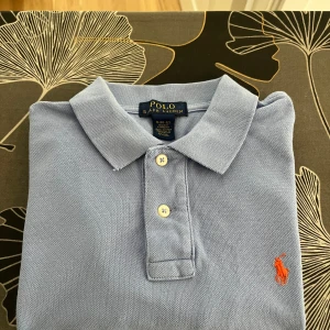 Ljusblå pikétröja från Polo Ralph Lauren - Klassisk ljusblå pikétröja från Polo Ralph Lauren med korta ärmar, krage och tre knappar framtill. Tröjan har den ikoniska orange broderade loggan på bröstet och är tillverkad i bomull för en skön och luftig känsla.