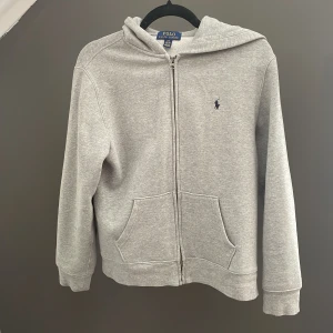 Grå hoodie från Polo Ralph Lauren - Klassisk grå hoodie från Polo Ralph Lauren med dragkedja framtill, huva och fickor. Broderad marinblå logga på bröstet. Mjuk bomullsblandning och ribbade muddar för extra komfort. Perfekt för chill dagar eller när du vill ha en clean look. Aldrig använd. Nypris 1200kr.