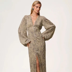 Adooreklänning zebra - Snygg långklänning med zebramönster i beige och grått. Djup v-ringning, långa puffärmar och knappar framtill hela vägen ner. Klänningen har en slits framtill som ger extra edge. Perfekt för dig som vill sticka ut med en trendig look.