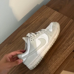 Nike Dunk Low i vitt och grått - Nike Dunk Low sneakers i vitt och ljusgrått med klassisk Swoosh-logga på sidan. Skon har snörning, rund tå och platt sula. Ovandelen är i skinn och syntet, perfekt för en clean och stilren look.
