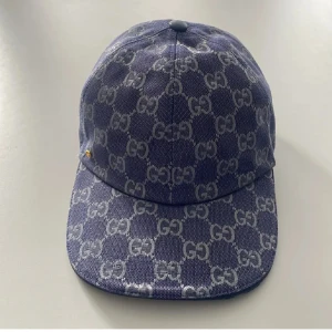 Blå Gucci keps med GG-mönster - Snygg blå keps från Gucci med ikoniskt GG-mönster i grått över hela kepsen. Klassisk böjd skärm och justerbar rem baktill. Tillverkad i bomullsmix med exklusiv känsla och diskret guldfärgad detalj på sidan. Perfekt för dig som vill sticka ut med lyxig streetstyle. Priset går att diskuteras 