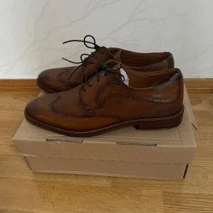 Bruna klassiska skinnloafers med snörning - Snygga bruna loafers i skinn med klassisk broguedetalj och snörning. Skorna har rund tå och låg klack, perfekt till en dressad look. Fina detaljer längs kanterna och stilren design som passar till många outfits.